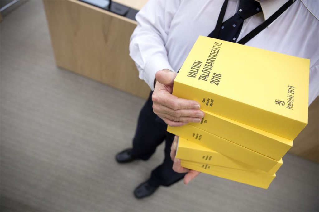 Henkilö kantamassa pinoa keltaisia kirjoja, joiden kannessa lukee "Valtion talousarvioesitys 2016".