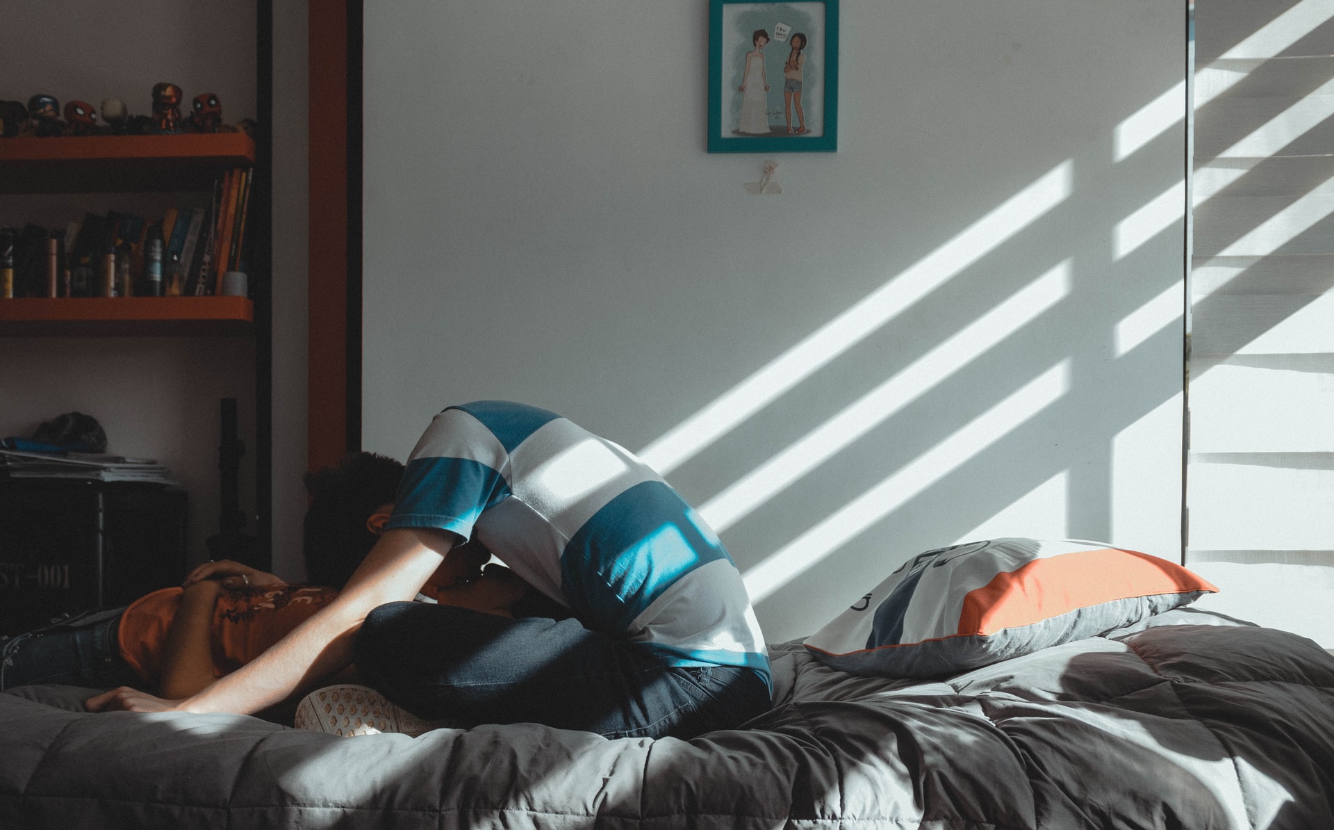 Sängyllä auringonvalossa istuva nuori henkilö kyyristyy suutelemaan toista
