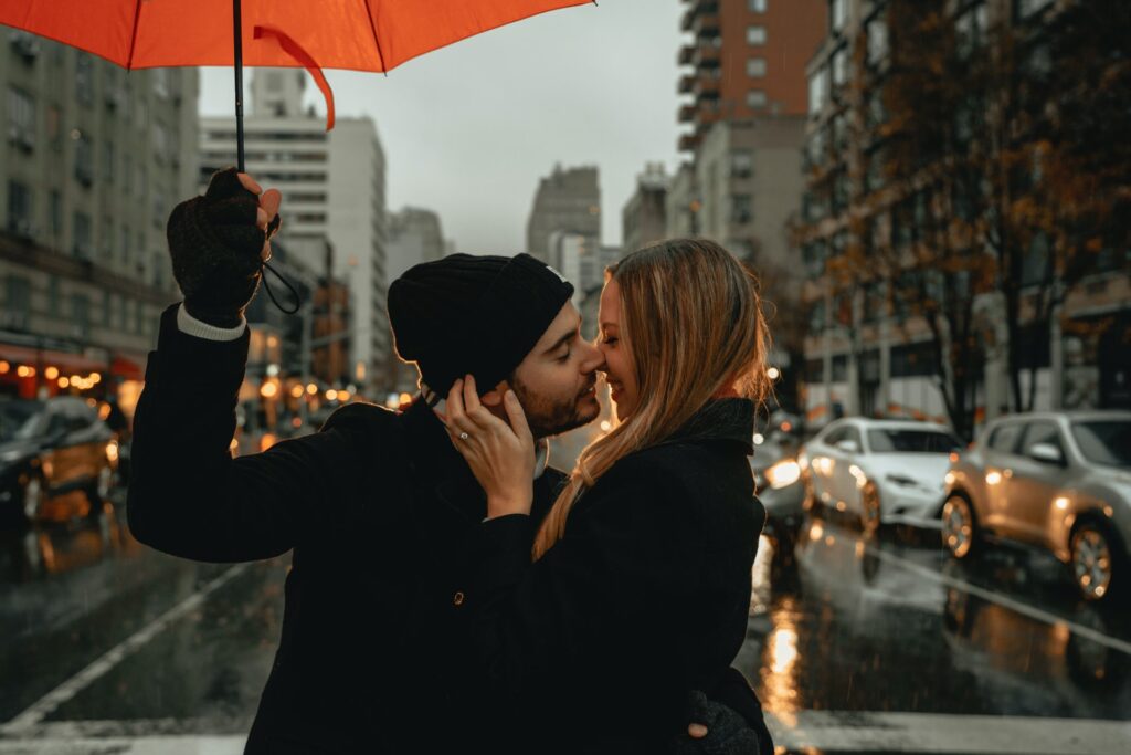 Kaksi henkilöä suutelee  ja hymyilee autotiellä sateenvarjon alla.