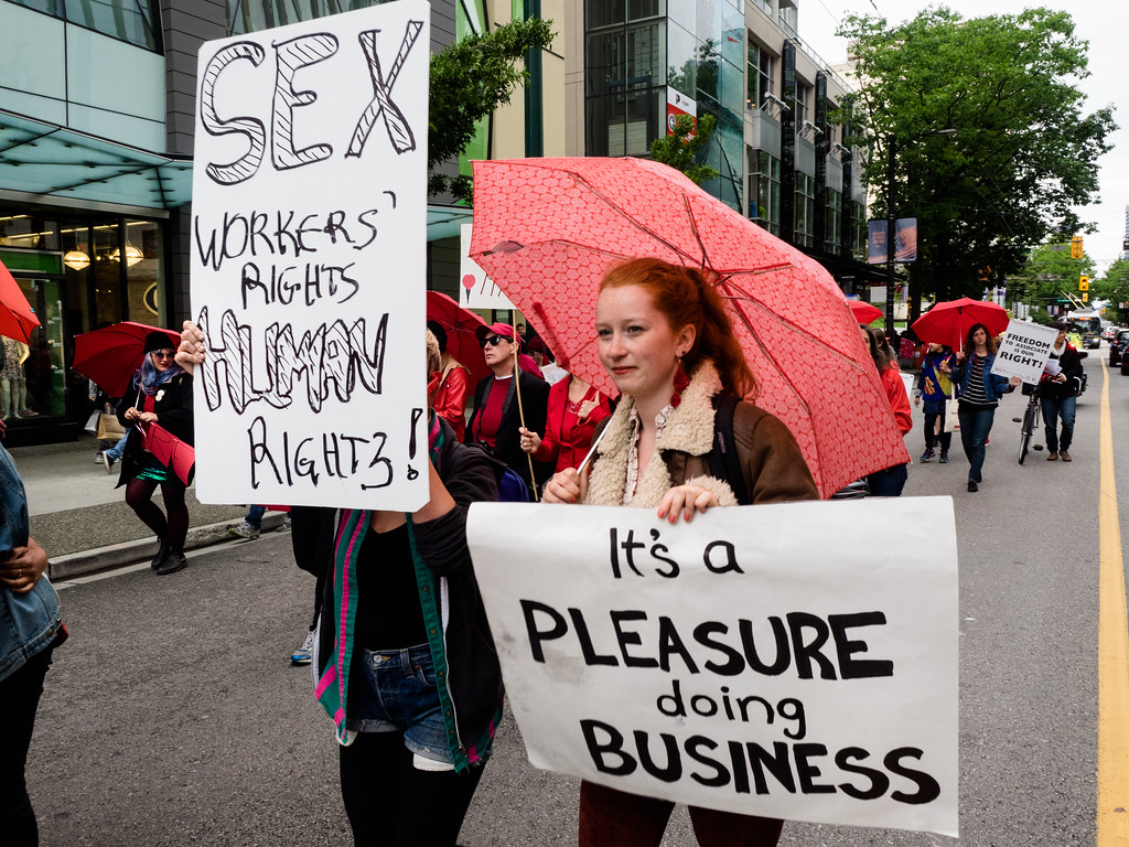 Punaisia sateenvarjoja kantavia ihmisiä mielenosoituksessa. Yksi heistä kantaa kylttiä, jossa lukee "Sex workers` rights human rights!". Hänen vieressään olevan kyltissä lukee "It's a pleasure doing business".
