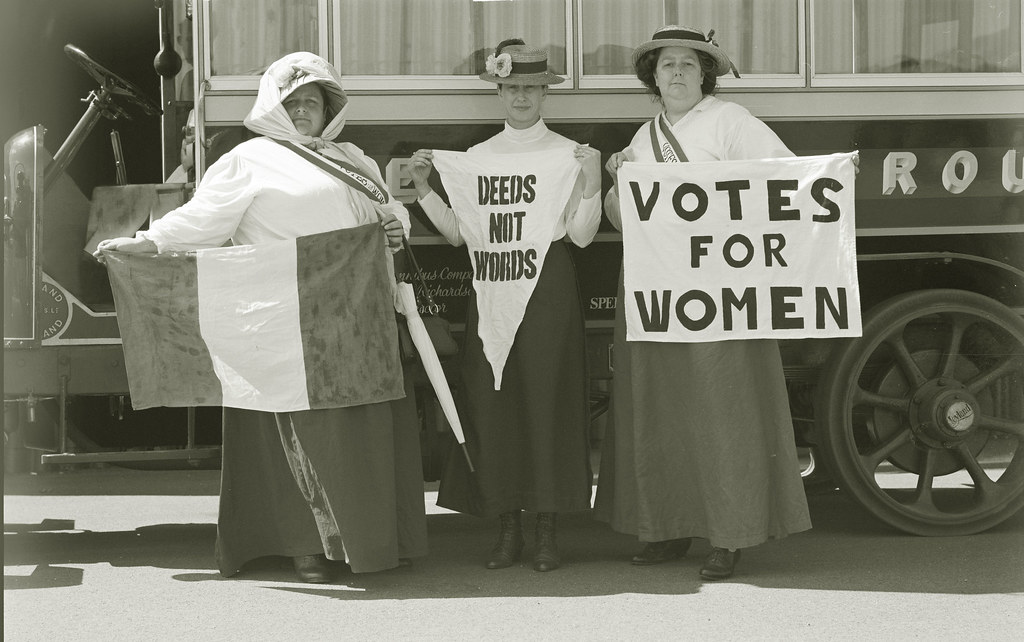 Naisasianaisia 1900-luvun alusta. He pitelevät kylttejä, joissa lukee "Deeds not words" ja "Votes for Women" eli toimintaa ei sanoja ja äänioikeus naisille.