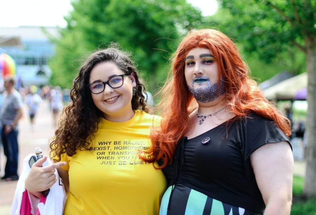 Kaksi henkilöä. Toisella on glitteriä parrassaan ja pitkät, punaiset hiukset. Toinen osoittaa paitaansa, jossa lukee: "Why be racist, sexist, homophobic or transphobic when you can just be quiet?".