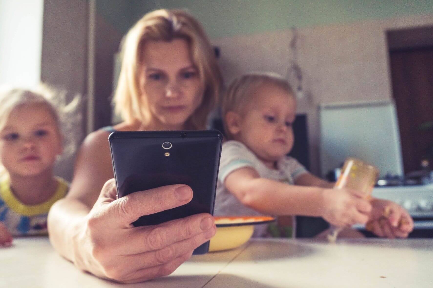 Äitihahmo käyttää älypuhelinta ruokapöydässä. Hänellä on sylissään yksi lapsi, ja toinen istuu vieressä.