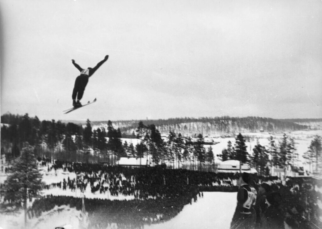 Mäkihyppääjä ilmassa kädet levällään Salpausselällä. Mustavalkoinen valokuva, 1960-luku. 