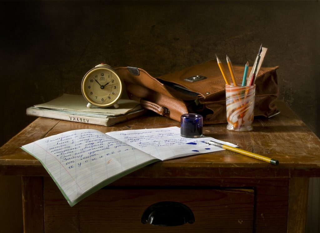 Puinen pöytä, jolla on vihko täynnä kaunokirjoitusta, mustekynä, kirja, herätyskello, koululaukku ja purkillinen kyniä. 