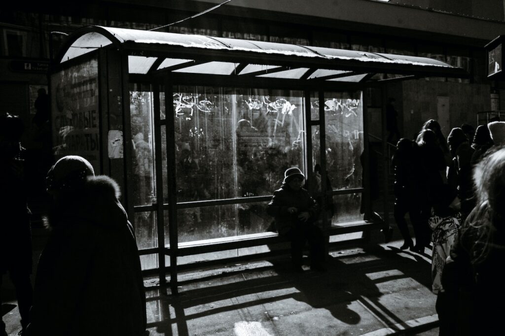 Mustavalkoinen kuva bussipysäkistä pimeällä. Pysäkillä istuvien ihmisten kasvot ovat varjossa.