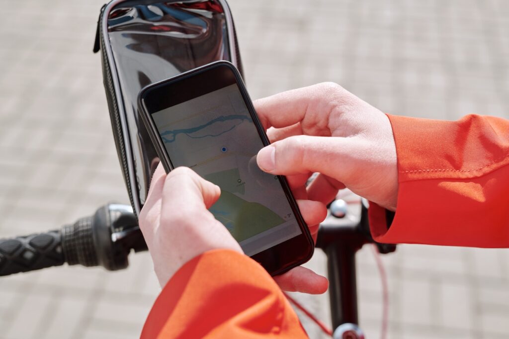 Kädet pitelemässä älypuhelinta, jossa on auki GPS-pohjainen karttasovellus. Taustalla näkyy pyörän ohjaustanko ja siihen kiinnitetty puhelinpidike.