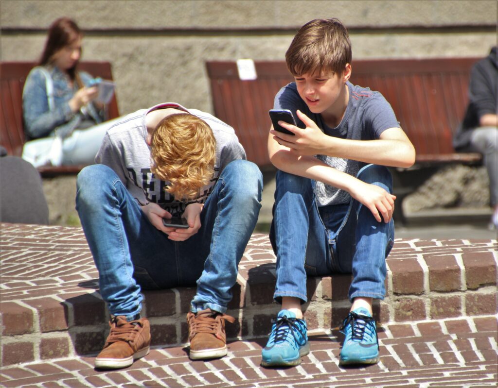 Kaksi teini-ikäistä poikaa katselee intensiivisesti puhelimiaan kiveyksellä. Taustalla näkyy nainen, joka istuu puhelin kädessä.