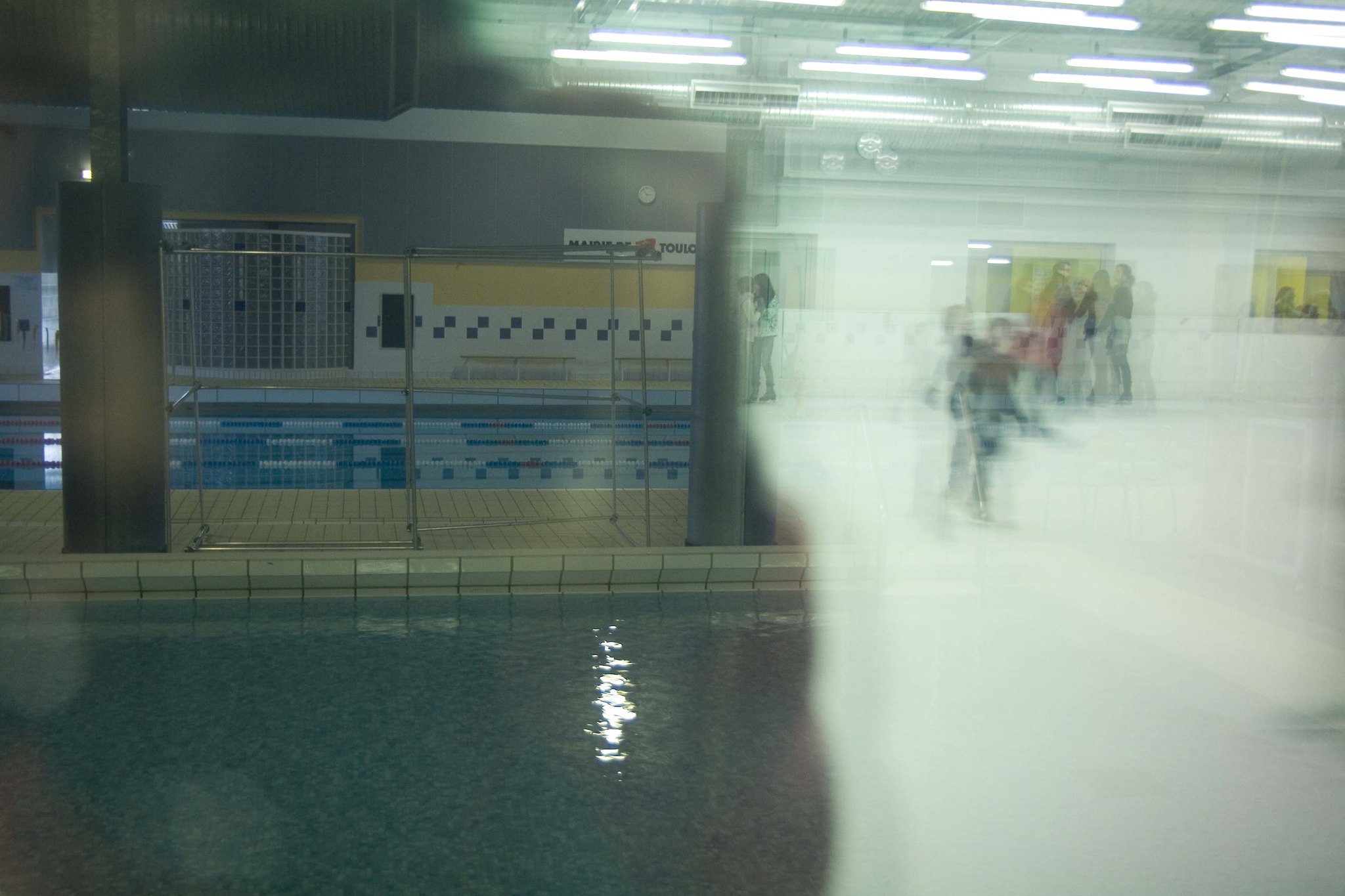 Sumea kuva uimahallista, joka on otettu ikkunan takaa. Kuvassa oikealla näkyy epätarkasti piirtyneitä hahmoja, kuvassa vasemmalla on uimahallin uimaosasto. 