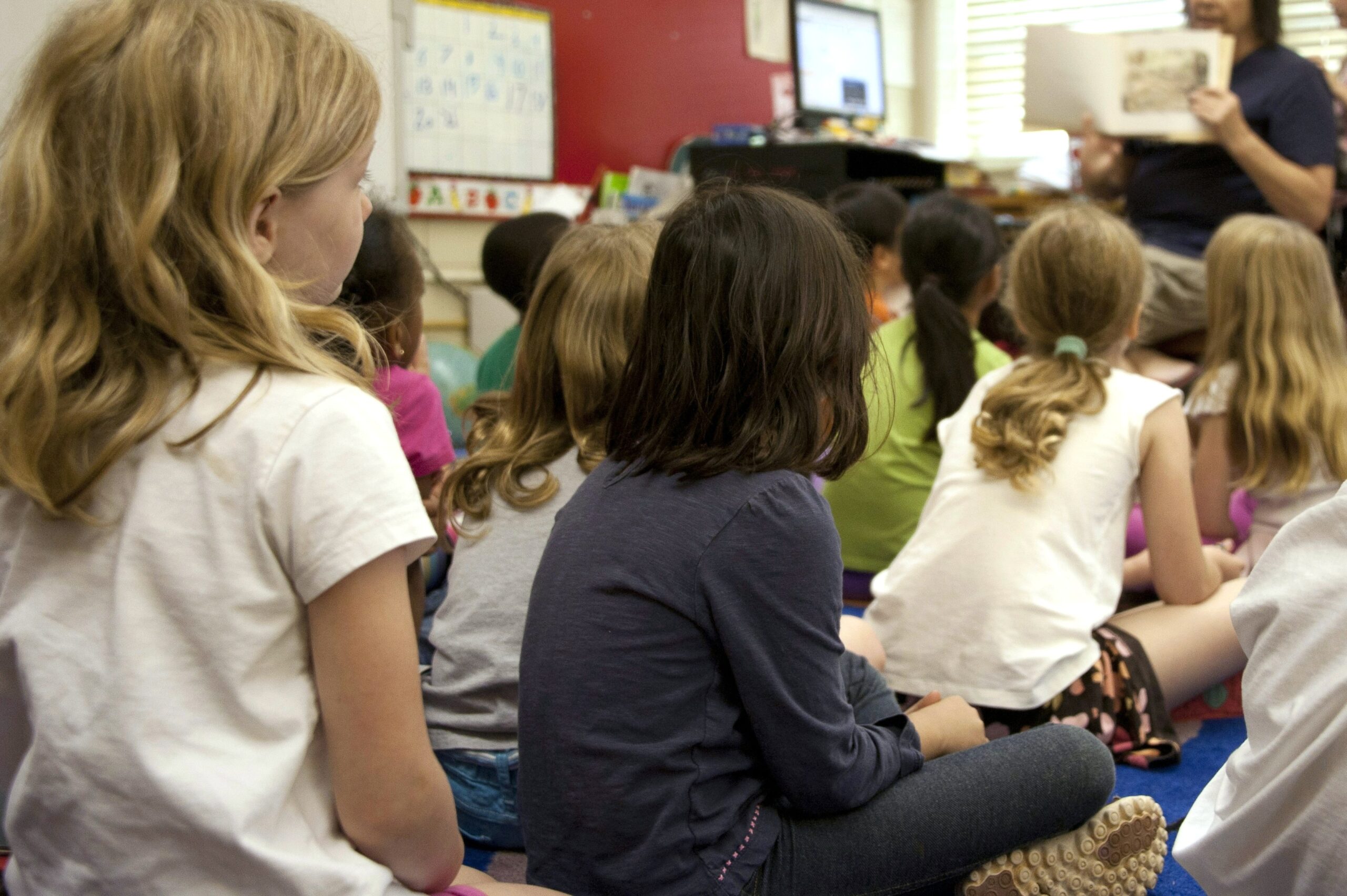 Ryhmä lapsia istuu lattialla lukkohuoneessa selin kameraan. Edessä aikuinen esittelee lapsille kirjaa.