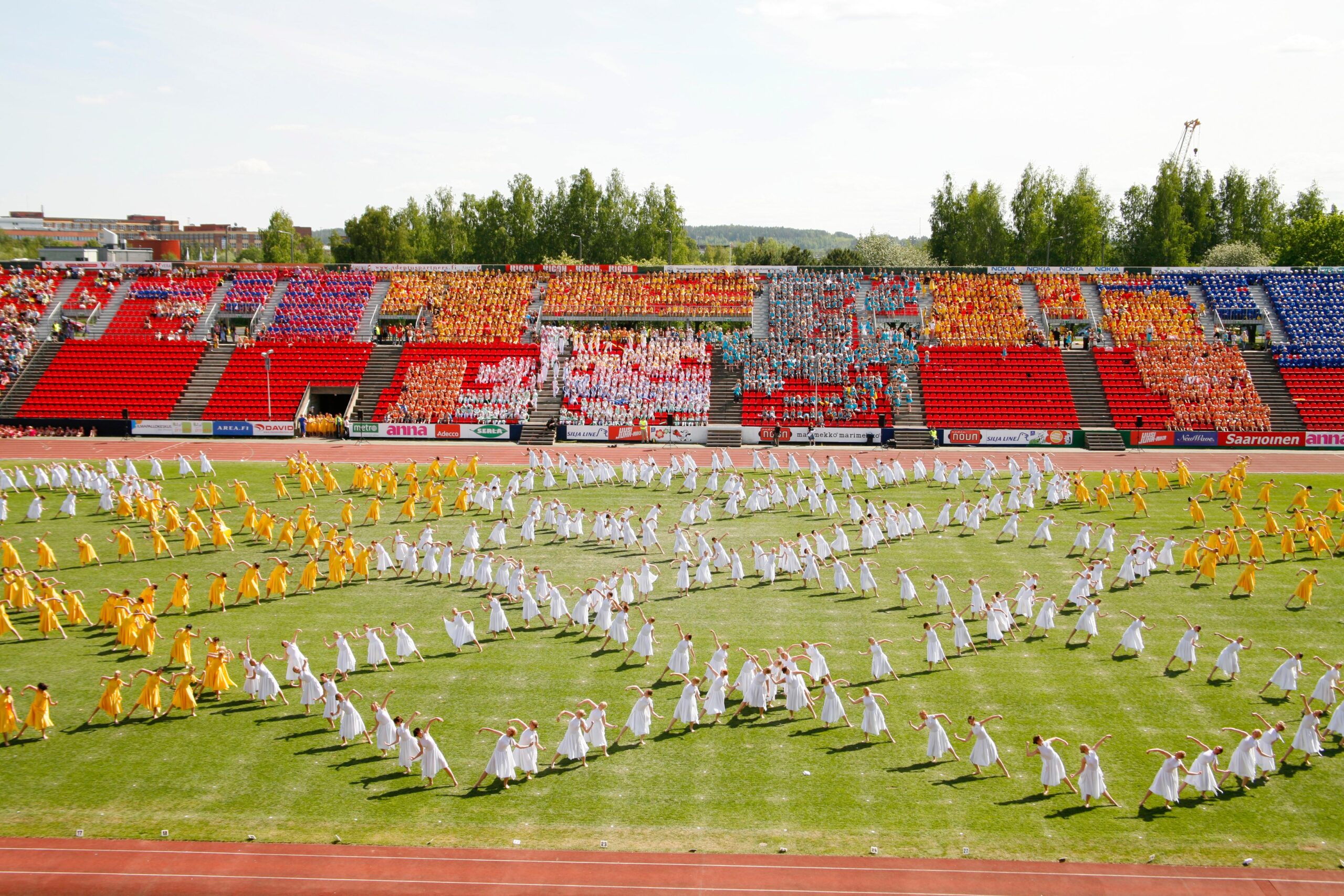 SUN SVOLI - voimistelun iloa suurtapahtuma Ratinan stadionilla Tampereella. Kuvassa monia voimistelijoita kentällä pukeutuneena valkoisiin ja keltaisiin mekkoihin. Voimistelijat muodostavat kentälle ruutumaisen muodostelman.