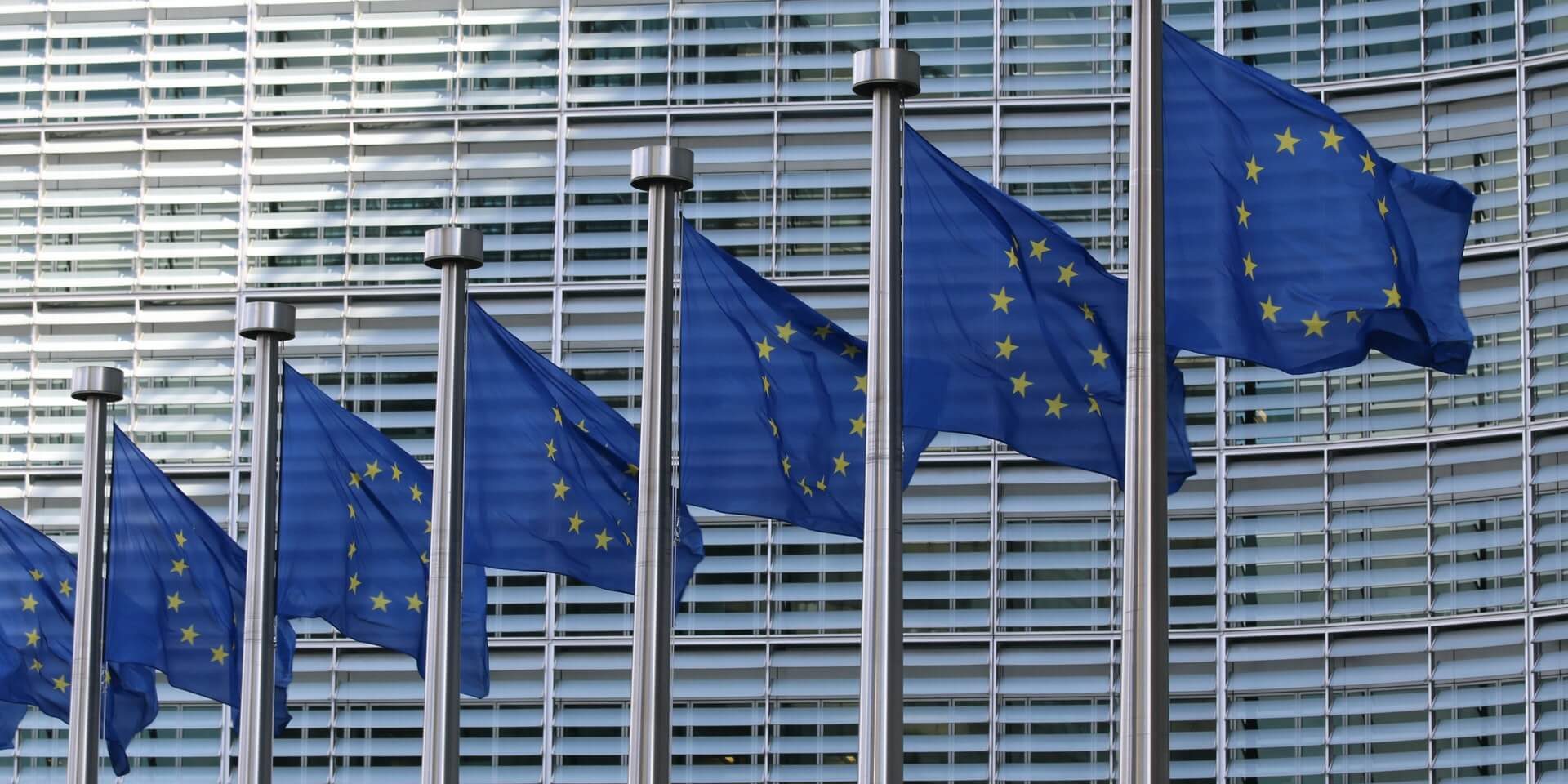 Seitsemän liehuvaa Euroopan unionin lippua harmaan rakennuksen edessä.