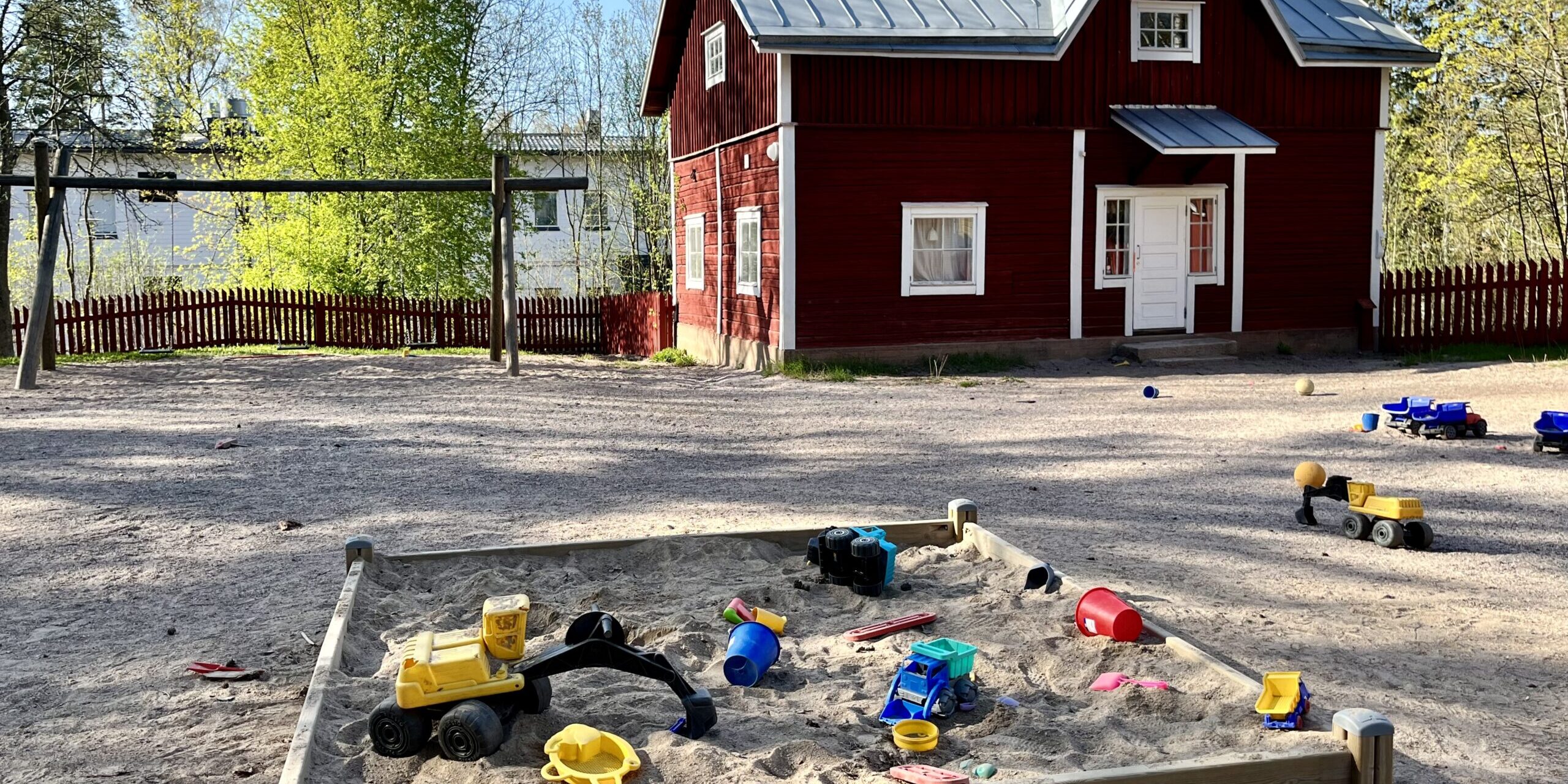 hkm.0bb0a6ea-d1b5-41ff-b92b-0da74dc0f077-0-original Kuvassa hiekkalaatikko punaisen puutalon edustalla. Pihalla on lasten leluja.