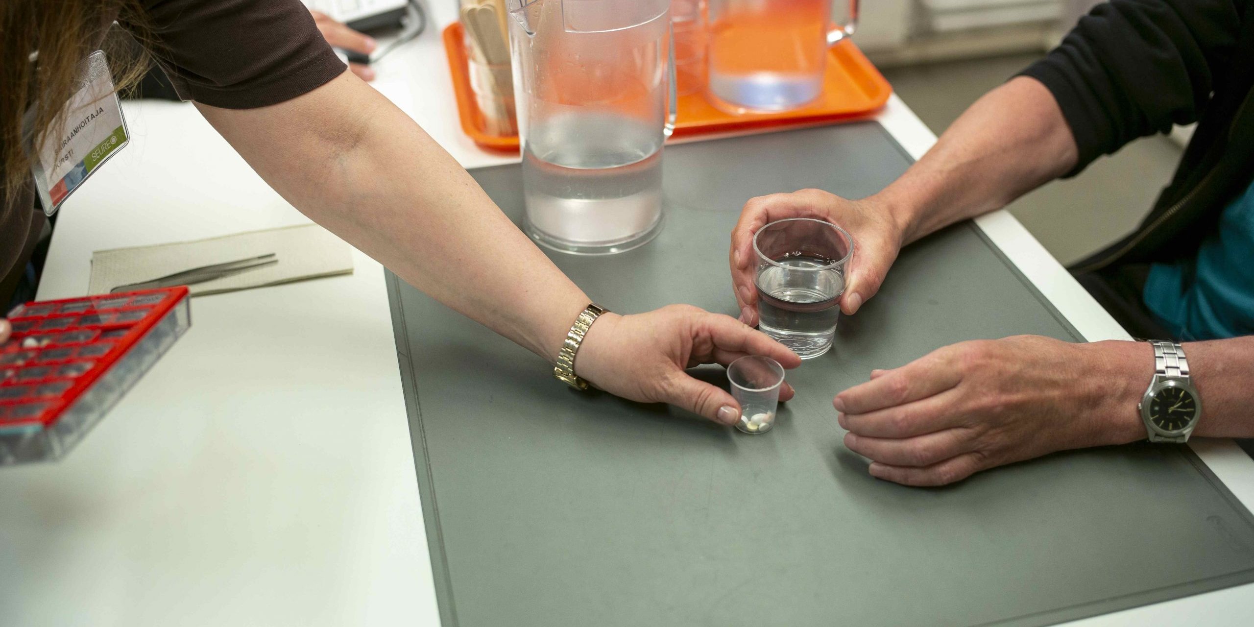 Hoitaja ojentaa vesilasia pitävälle ihmiselle lääkkeet pöydän ääressä