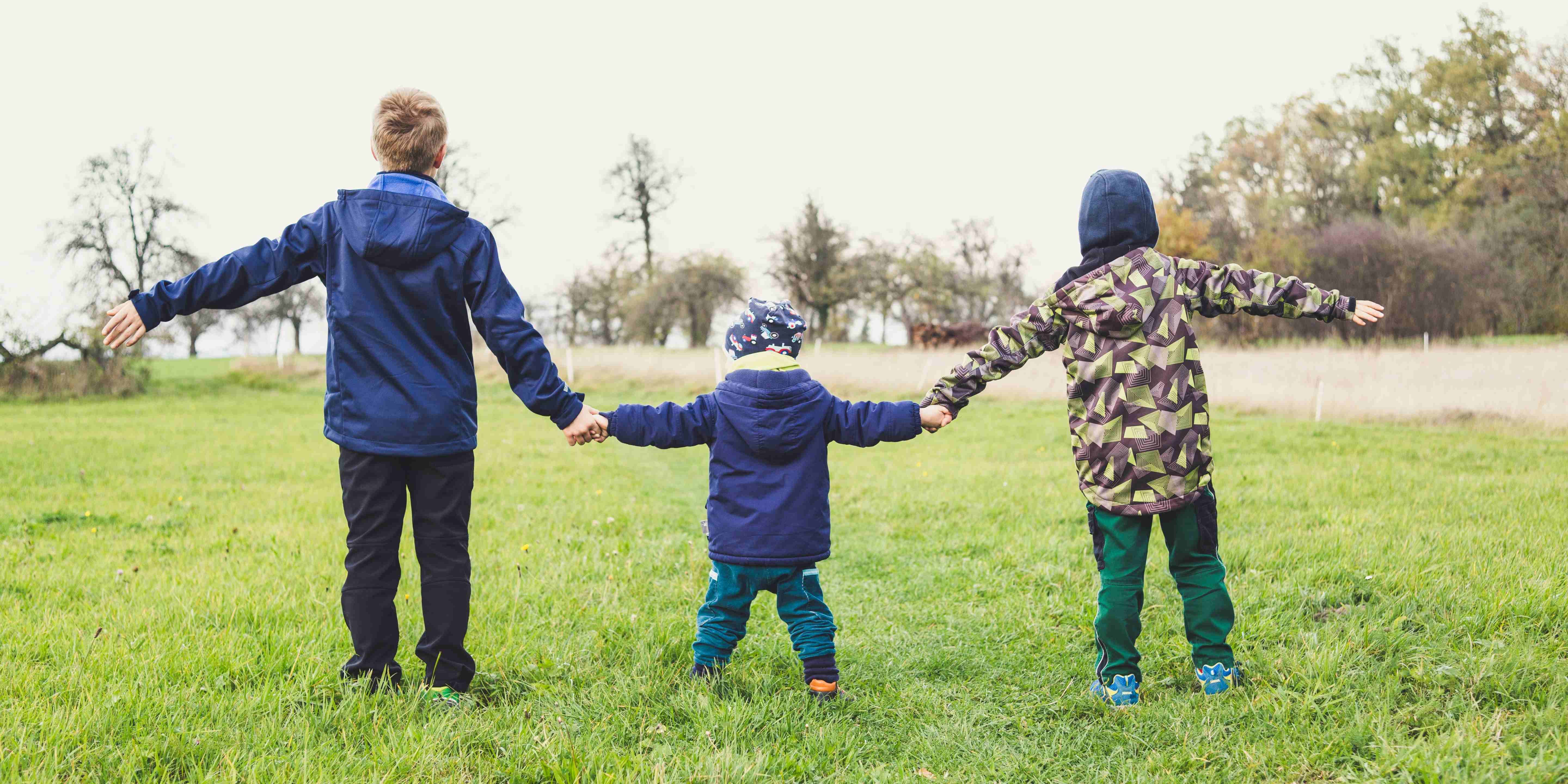 Kolme eri-ikäistä lasta pitelee toisiaan kädestä nurmikolla.