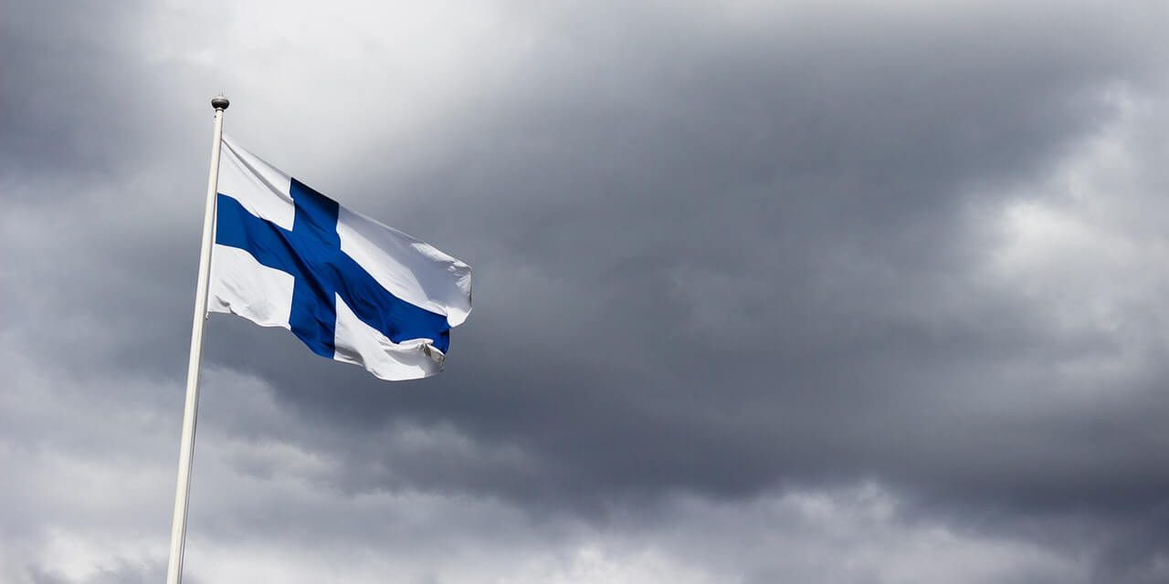 Suomen siniristilippu liehumassa tummanharmaata pilvistä taivasta vasten.