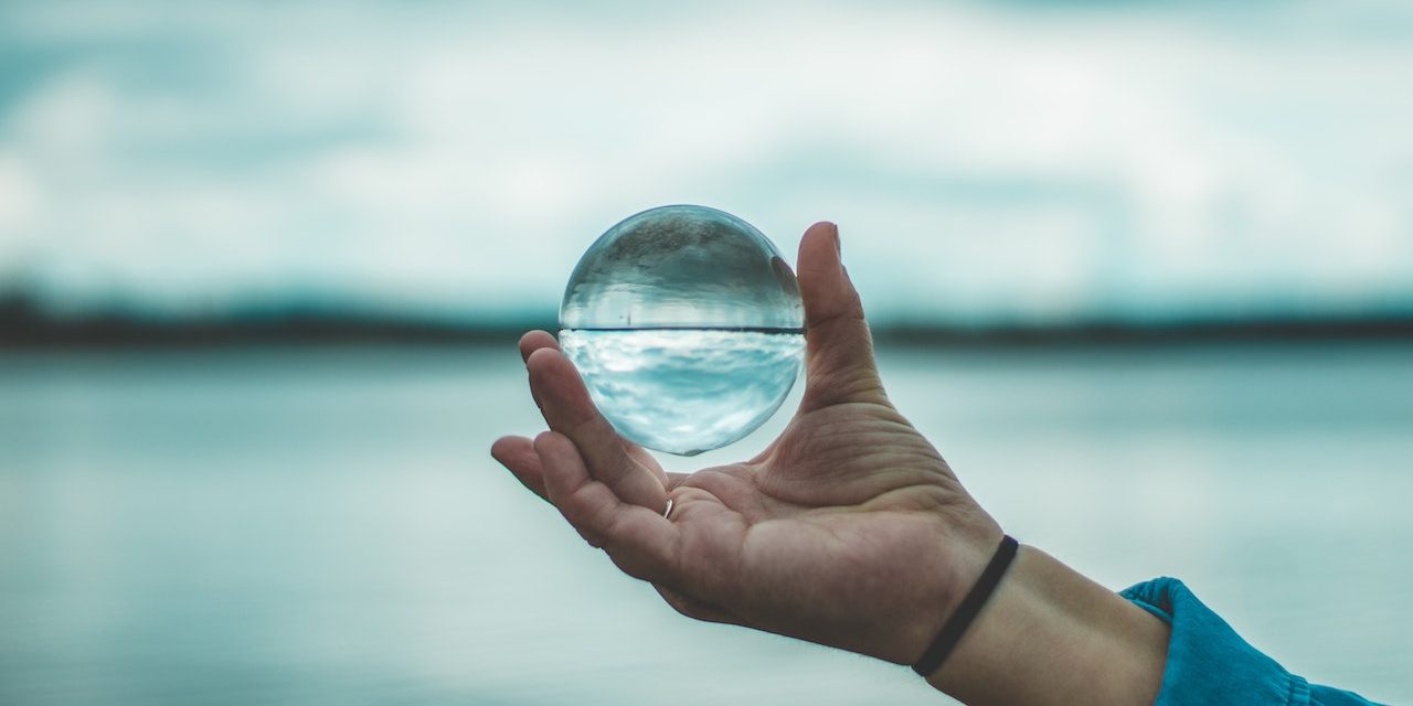 Henkilö pitää kädessään kristallipalloa. Taustalla veden aava, joka näkyy pallon sisällä ylösalaisin.