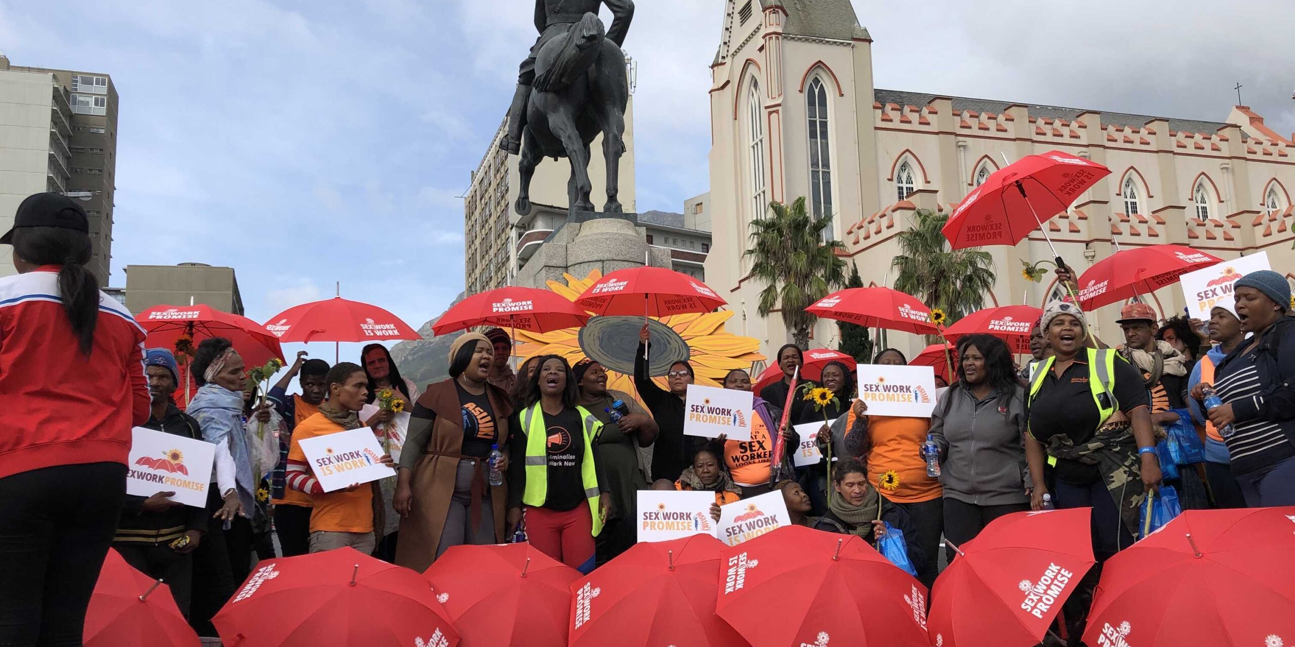 Henkilöitä seksityötä puolustavassa mielenosoituksessa. Monella on punainen sateenvarjo, jossa lukee "Sex work is work" ja "Sex Work Promise".